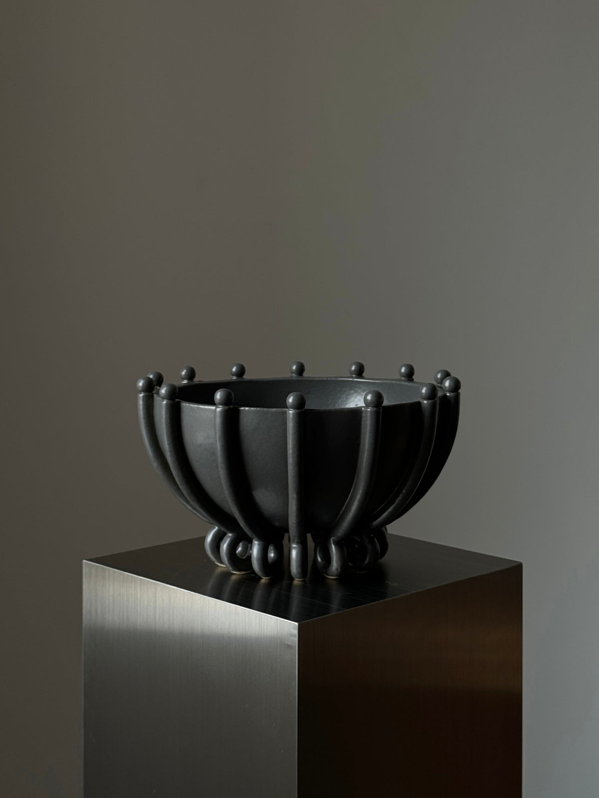 Vessel No. V by Devin Wilde is a sculptural black bowl featuring curved rib-like segments and small spherical accents, displayed on a metallic pedestal against a plain gray background.
