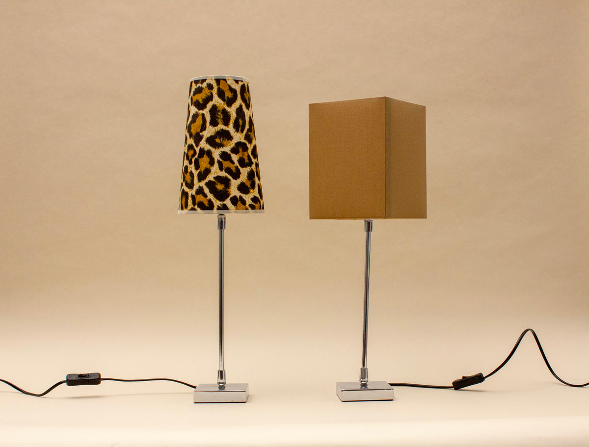 Two SÒLER ZAFRA TABLE LAMPs with chrome stands sit side by side. The left lamp has a bold leopard print shade; the right has a plain, square beige shade. Both cords are visible.
