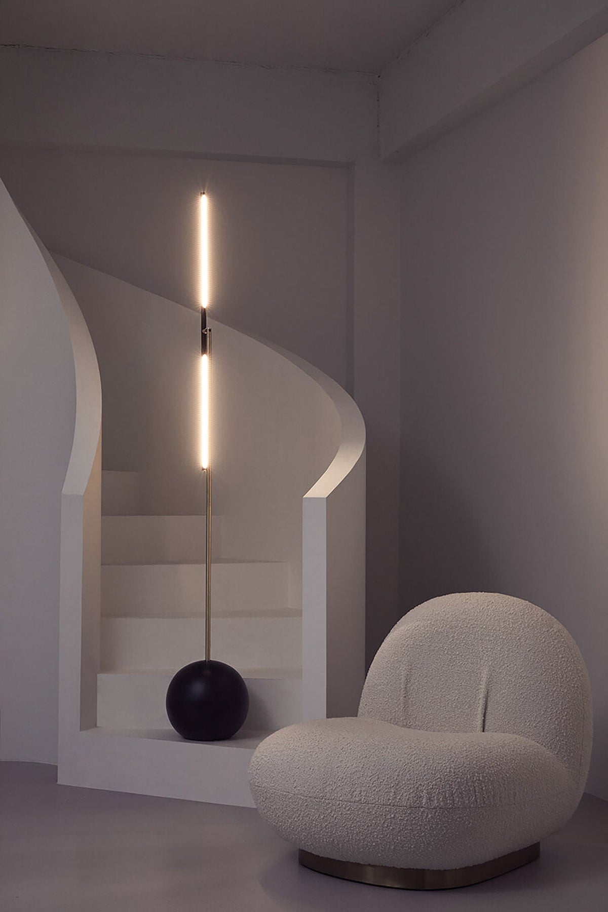 A modern, minimalist room with a curved white staircase, sculpture-like lighting, and a plush rounded lounge chair. The space features the 'T-Lamp' Floor Lamp by Heako Studio, casting soft light for a serene ambiance.