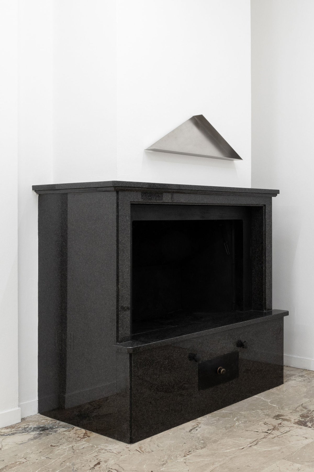 A modern black granite fireplace set against a white wall features "The Front - Shelf in Metal" by COSEINCORSO above, with beige marbled tiles and an architectural shelf adding sculptural decor to the minimalist look.