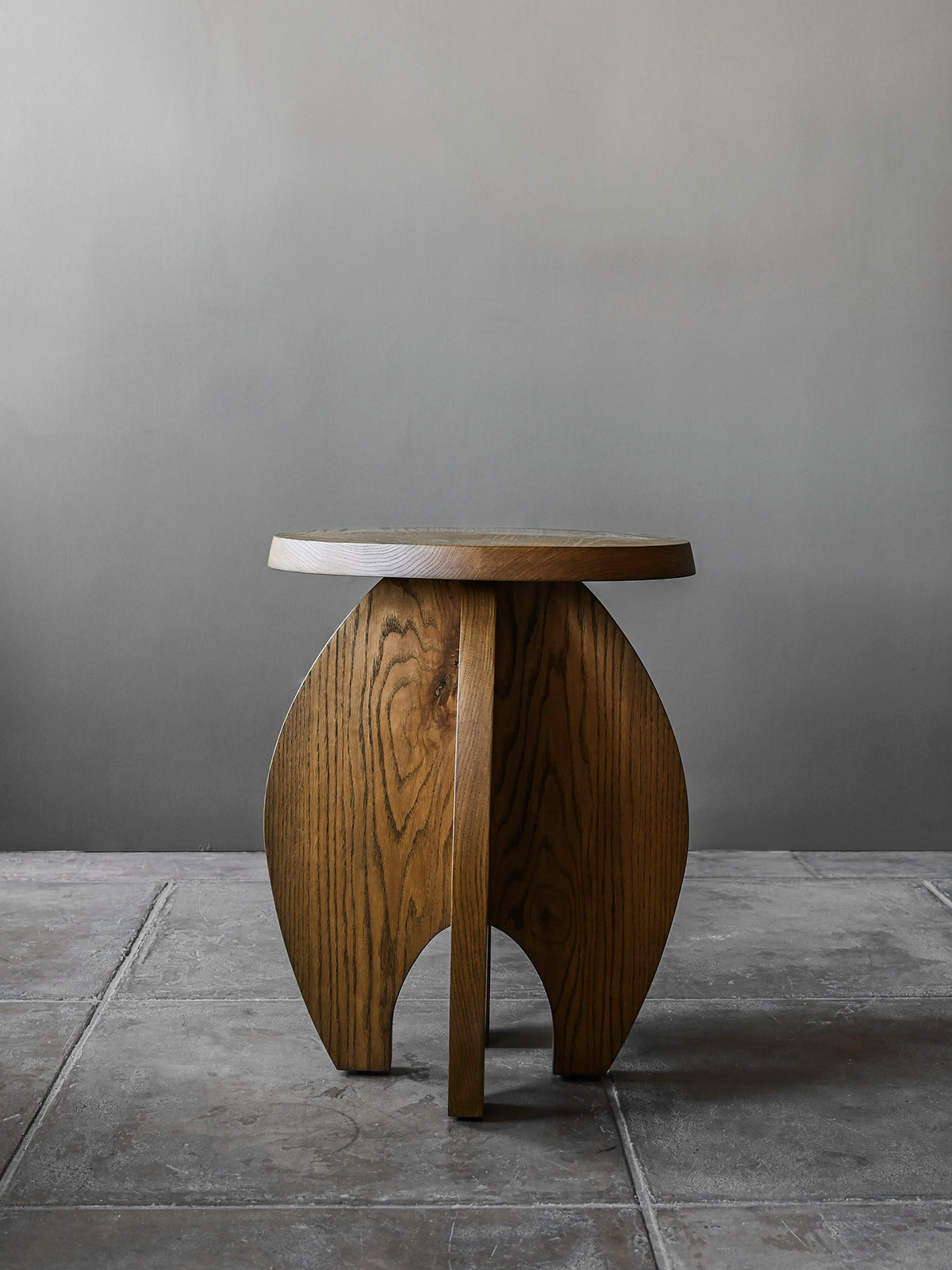 The 'Fusée' Stained Oak Side Table by Studio OSKLO features a small, round top and a rocket-shaped base with visible wood grain. It stands on a gray tiled floor against a plain gray wall.