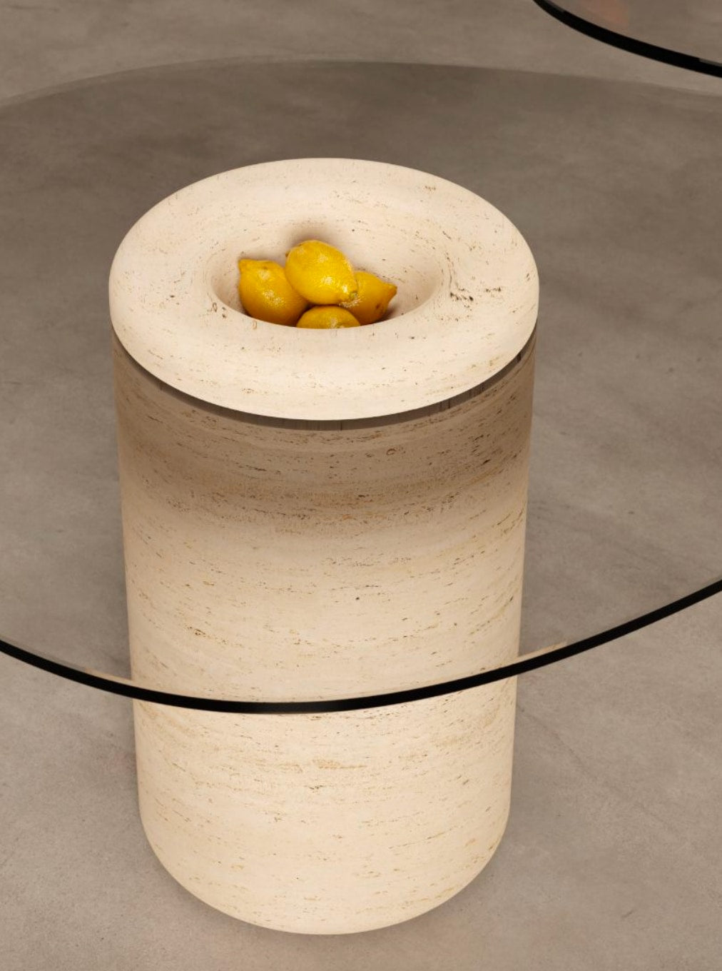 A cylindrical stone pedestal with a recessed top holds yellow lemons. Its organic form echoes the BURGIO. UUFO 01 Dining Table, which is partly visible on the smooth gray floor in the foreground.