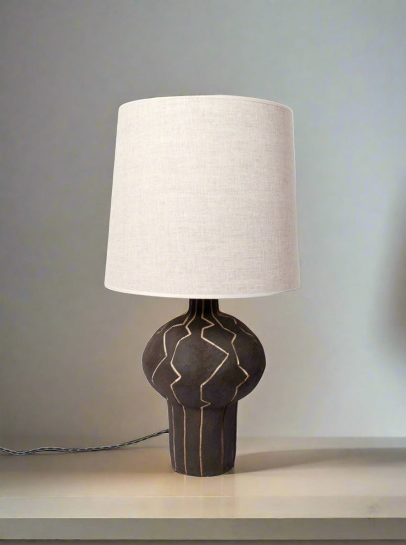 The "Lanzarote" Lamp by Barracuda Interiors features a black ceramic base with geometric lines and a large off-white fabric shade, providing customizable lighting on light surfaces in neutral spaces.