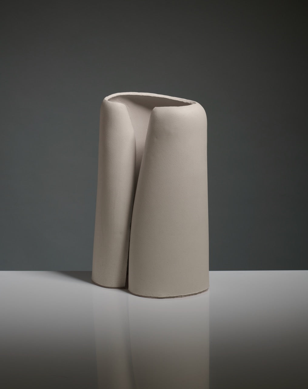The Snail Table Lamp by Katia Langeard features a minimalist, abstract white ceramic design with smooth curves and a top opening, shown on a reflective surface against a dark gray background.