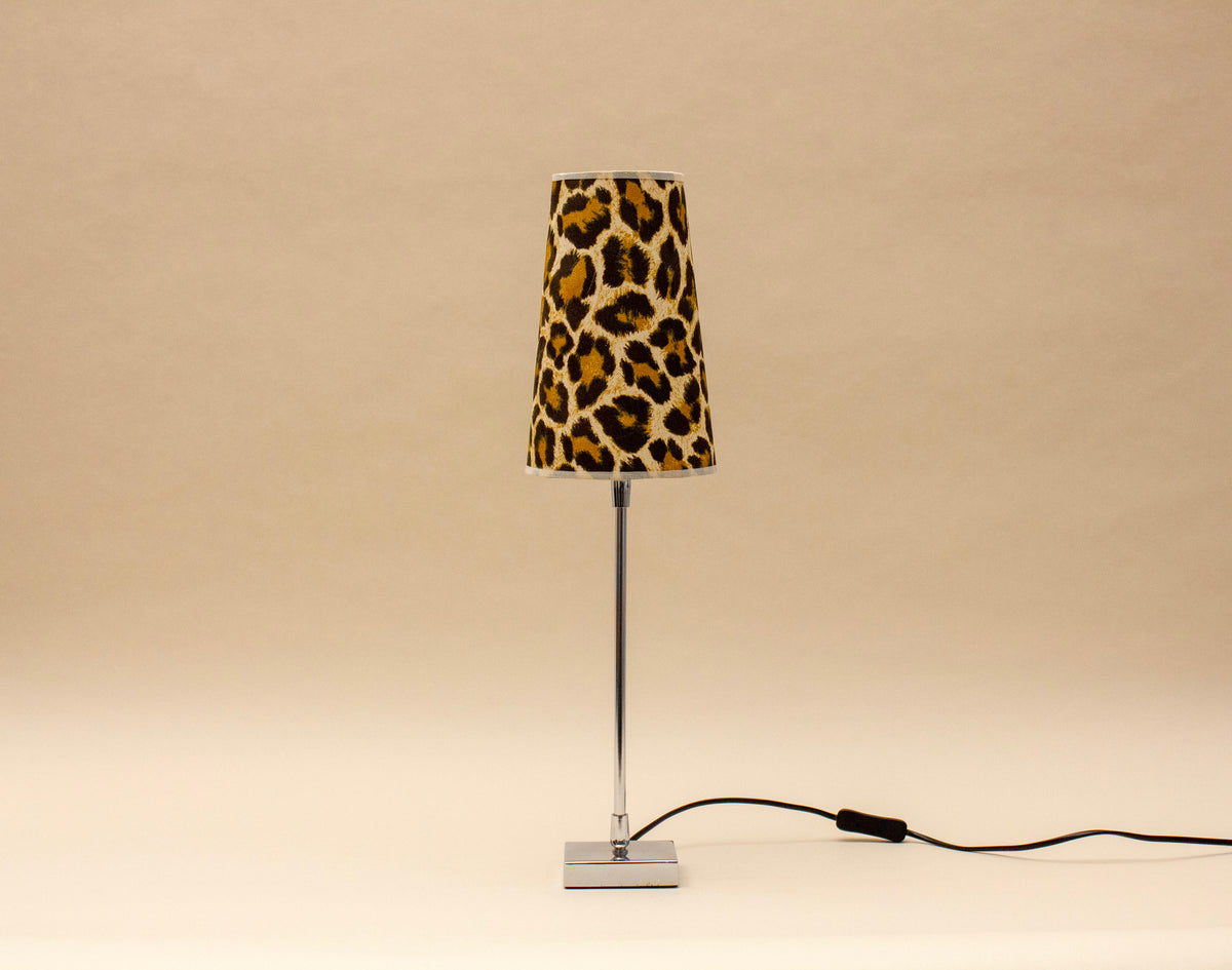 The SÒLER ZAFRA TABLE LAMP features a chrome base and a bold leopard print shade, resting on a neutral surface with its black power cord exposed. The plain beige background highlights its modern vintage design.