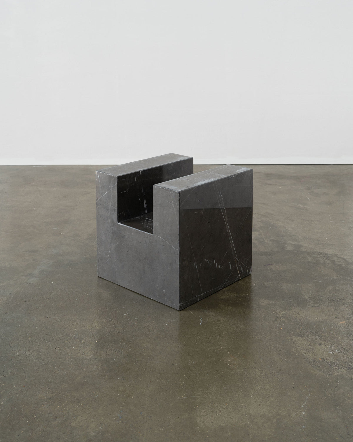 The Tom Bogle Postmodern Solid Marble Stool / Side Table features minimalist, rectangular lines and a dark marble finish, exuding elegance as it sits on a polished concrete floor against a plain wall.