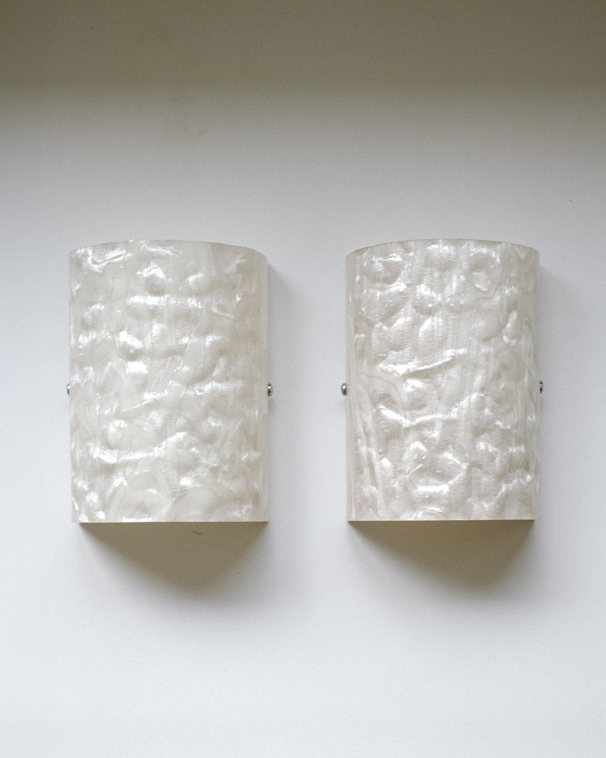 A Set of 2 Mother-of-pearl Wall Lamps by septembre studios, featuring a wavy, hammered texture and semi-cylindrical white design reminiscent of 1980s style, are mounted side by side on a light-colored wall.