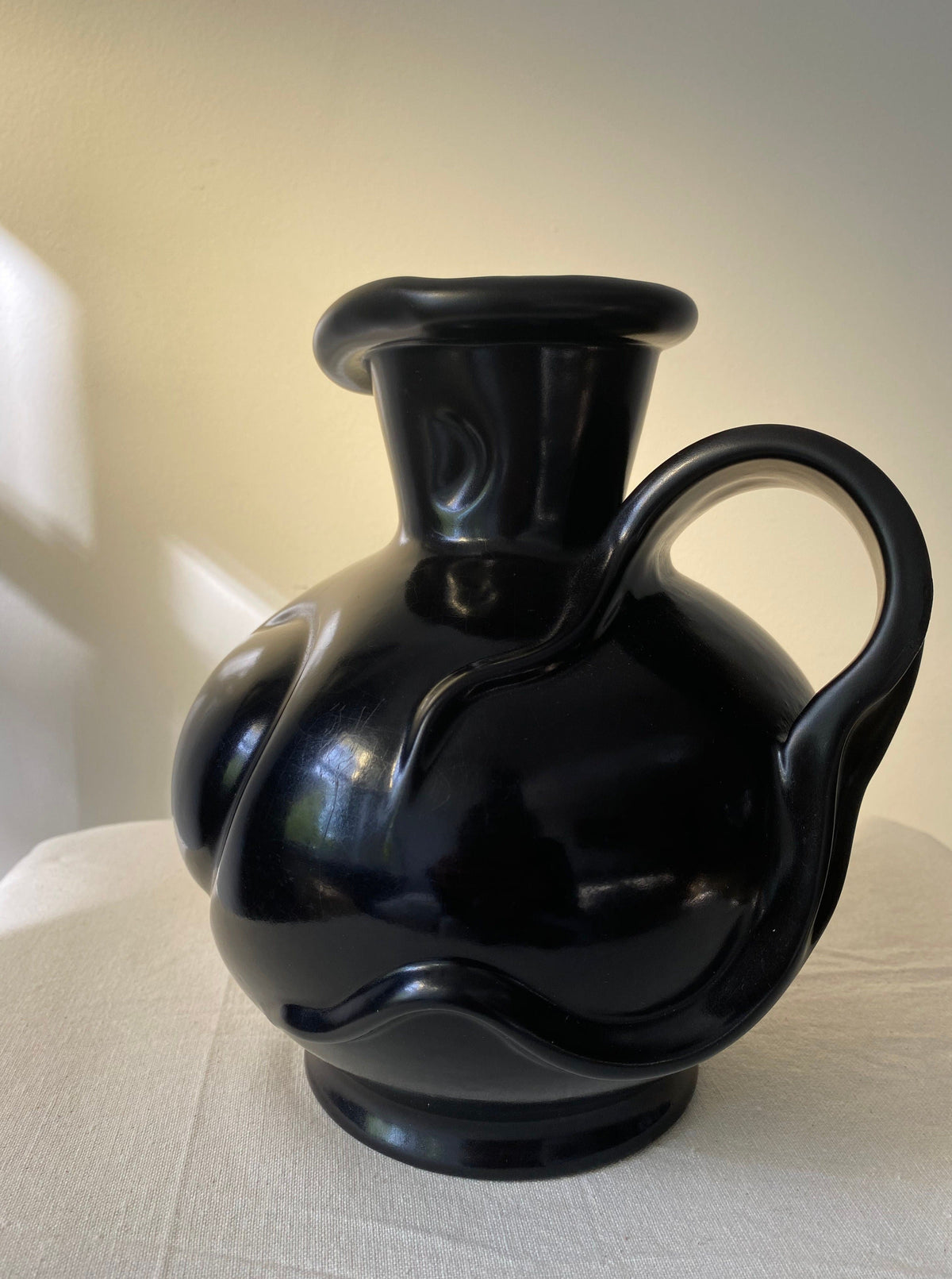 The Art Deco Vase by Les Objets Amsterdam, featuring a glossy black finish and wavy lines, rests on a cream cloth against a softly lit beige background.