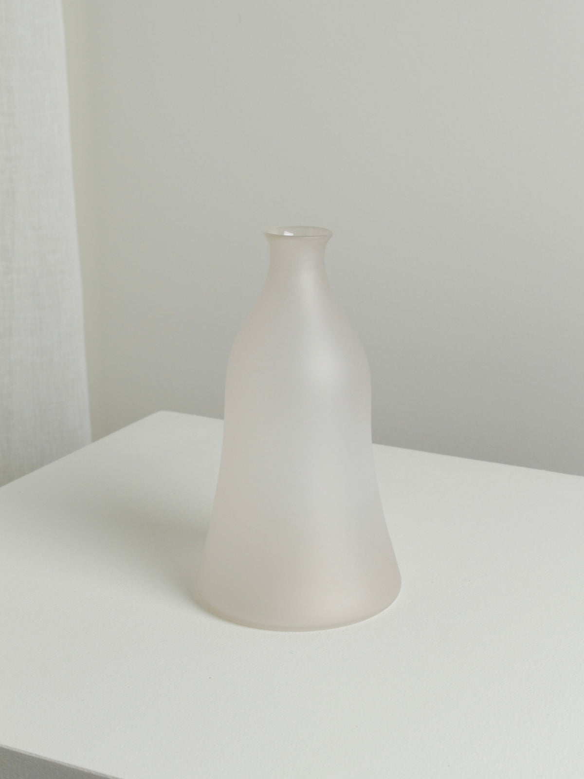 The Medium Vase in Moon by Verre d'Onge, featuring frosted glass with a narrow neck and wide base, rests on a white surface against a light-colored wall in a minimalist setting.