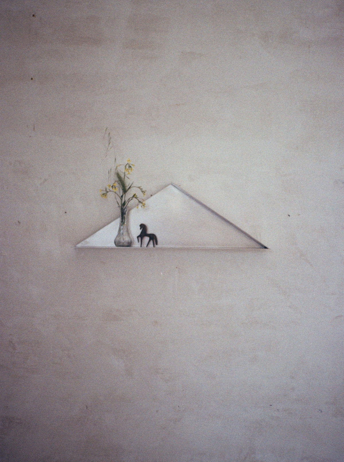 The Front - Shelf in Metal by COSEINCORSO, a minimalist triangular shelf, displays a glass vase with yellow flowers and a small black horse figurine against a light wall, accented by a stainless steel decorative detail.