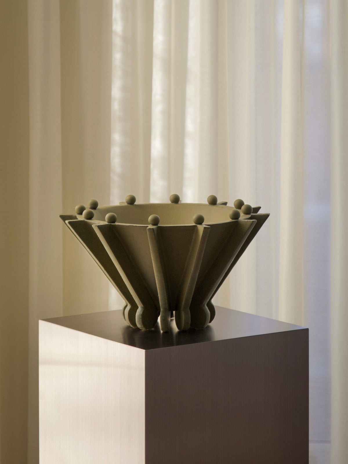 Vessel No. II by Devin Wilde, an olive-green ceramic bowl with geometric, flared sides and small spherical rim accents, sits on a square pedestal before sheer cream curtains.