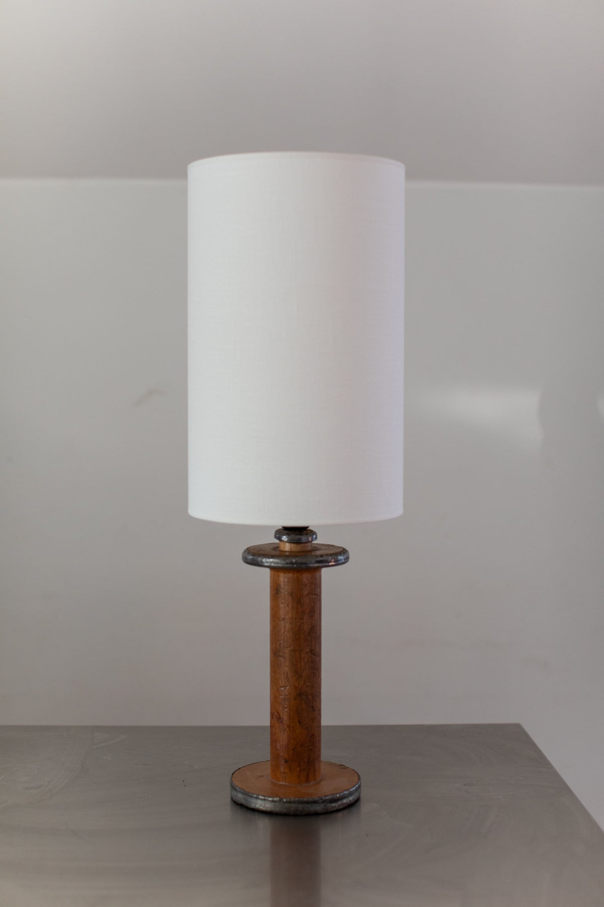 The Large Mid-Century Lamp by Out For Lunch, featuring a cylindrical white lampshade and a wooden handle base, stands on a smooth, gray surface. The background is plain and minimalistic, drawing attention to the lamp.