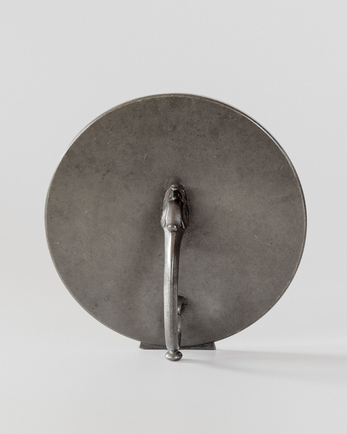 The Art Deco Metal Mirror by Spigel, featuring a round metal design with a central handle on the back, is reminiscent of a vintage shield. Displayed against a neutral gray background, it boasts a matte finish and simple design.