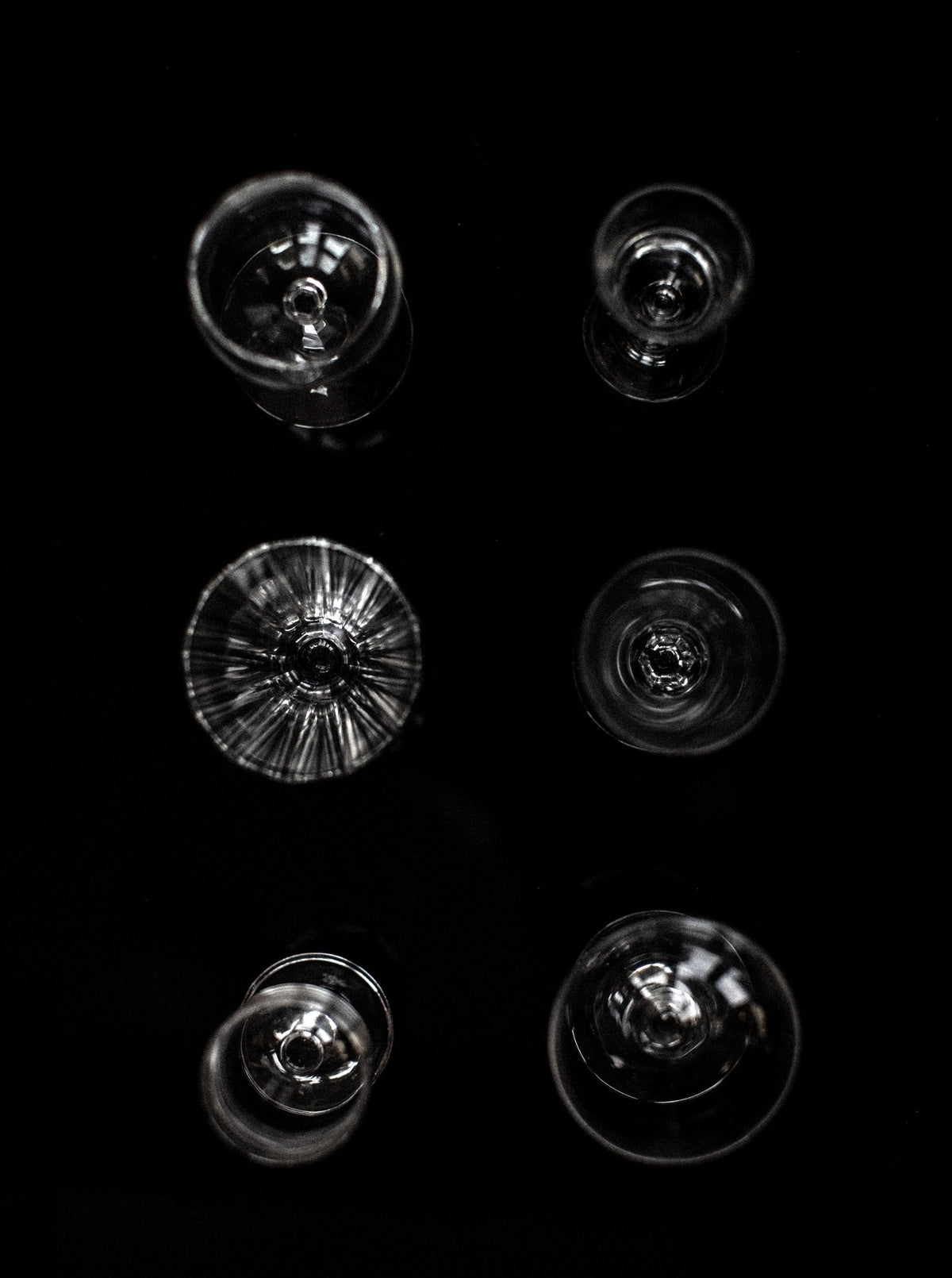 Overhead view of the Out For Lunch Set of Six Mix-Matched Glasses, artfully arranged on a black surface. Each glass is uniquely shaped and clear, with light gracefully reflecting off their rims and bases, offering a sophisticated drinking experience.