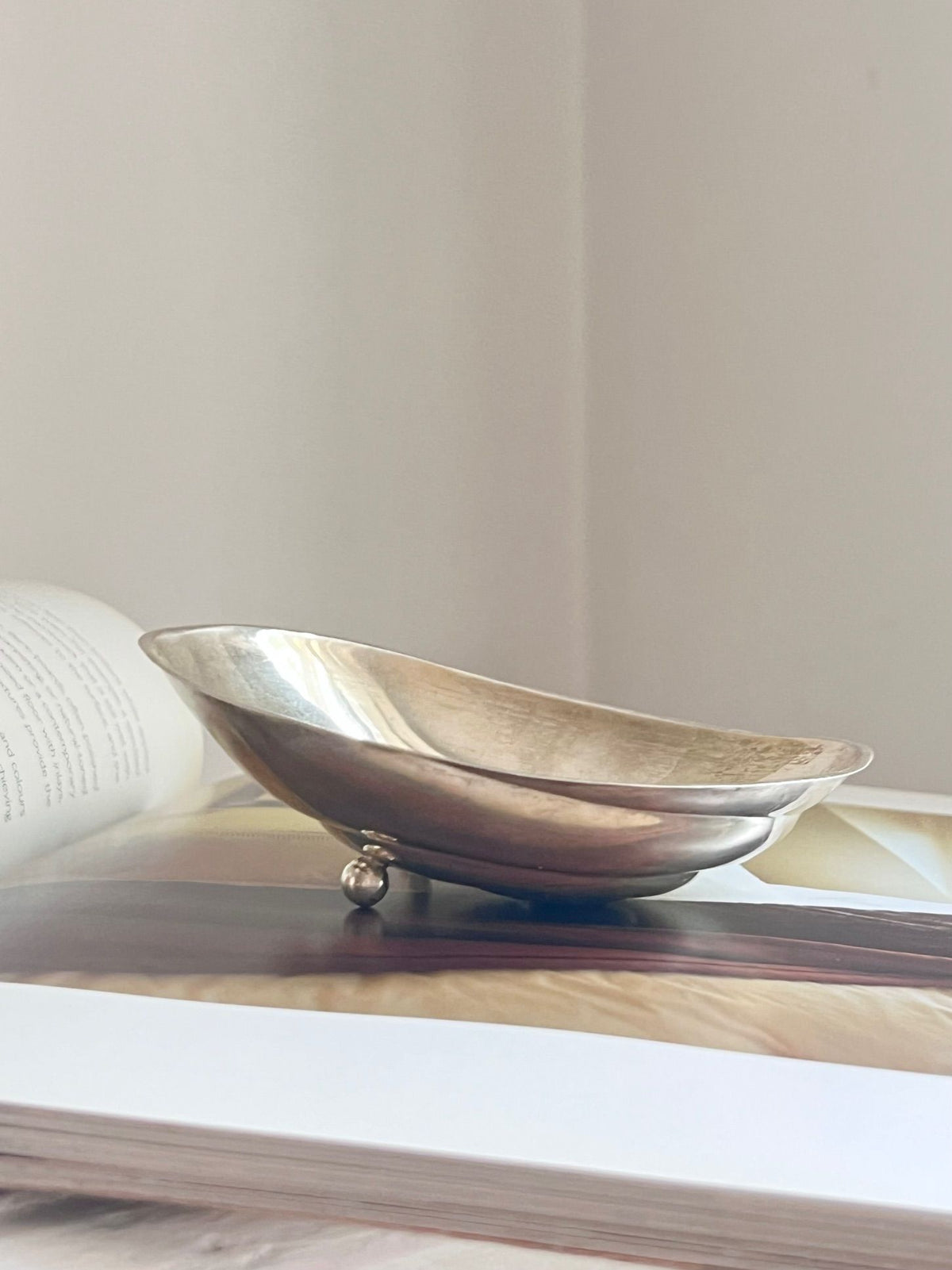 A Rare Sterling Silver Midcentury Vide-Poches by Médecine with three small legs is placed atop an open book. The background is minimalist and light-colored, giving the image a clean and serene atmosphere, reminiscent of a midcentury piece.