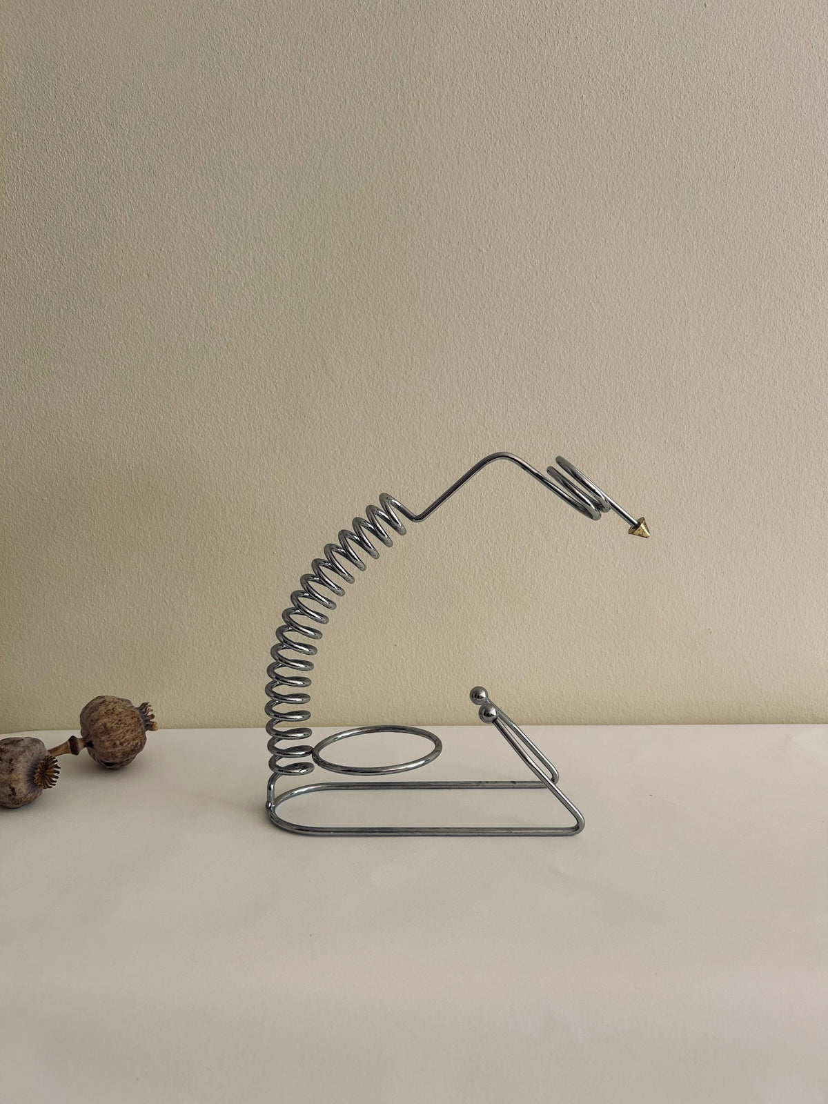 A sleek metal spiral resembling a coiled spring with a triangular tip, part of the minimalist design of Flock Studio's Postmodern Wine Holder, stands against a plain beige background. Two dried seed pods lie on the left side of the white surface, blending art with functionality.