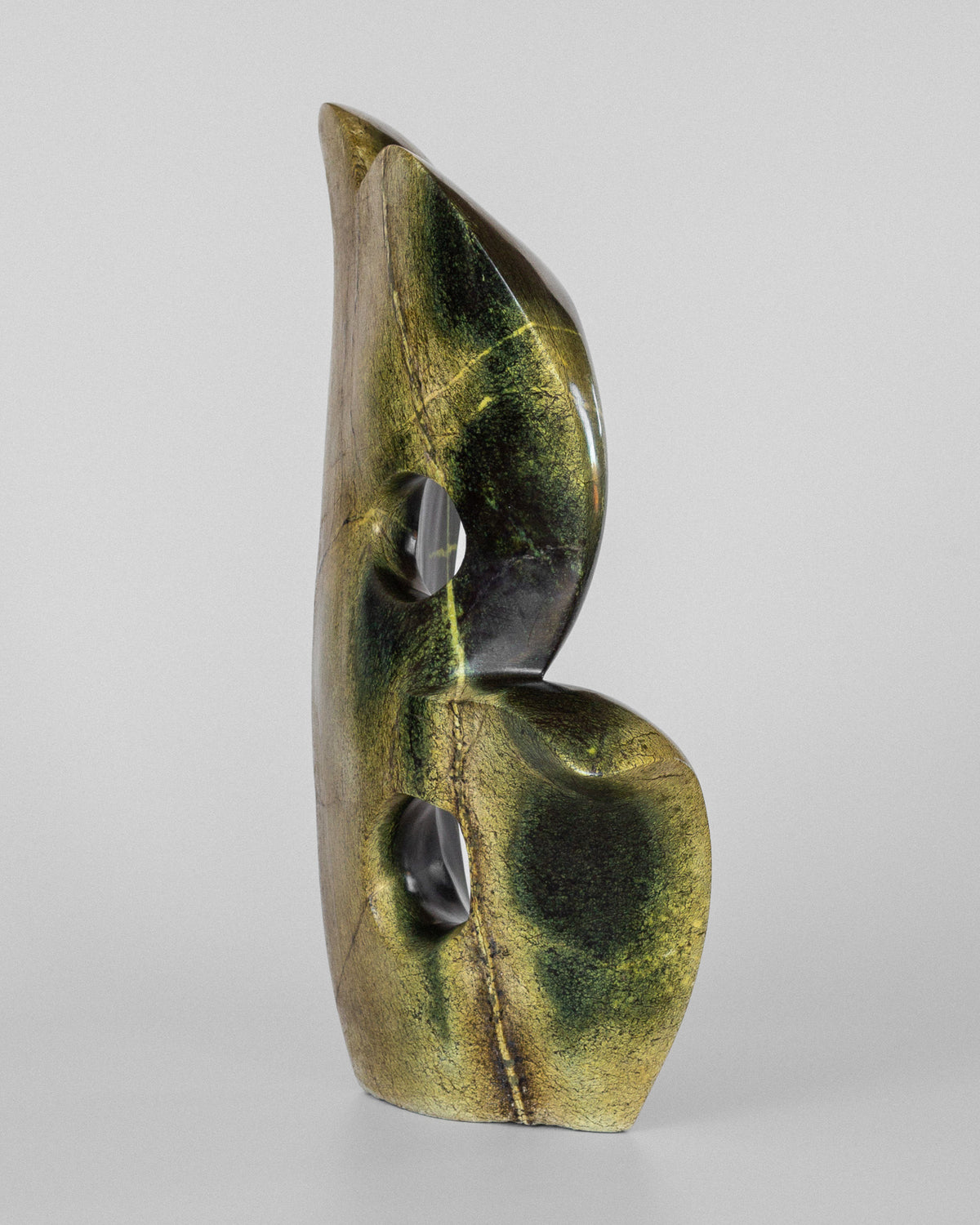The Abstract Stone Sculpture II by Spigel features smooth, curved surfaces with two circular openings and a green and gold marbled texture like leopard stone, all set against a plain gray background.