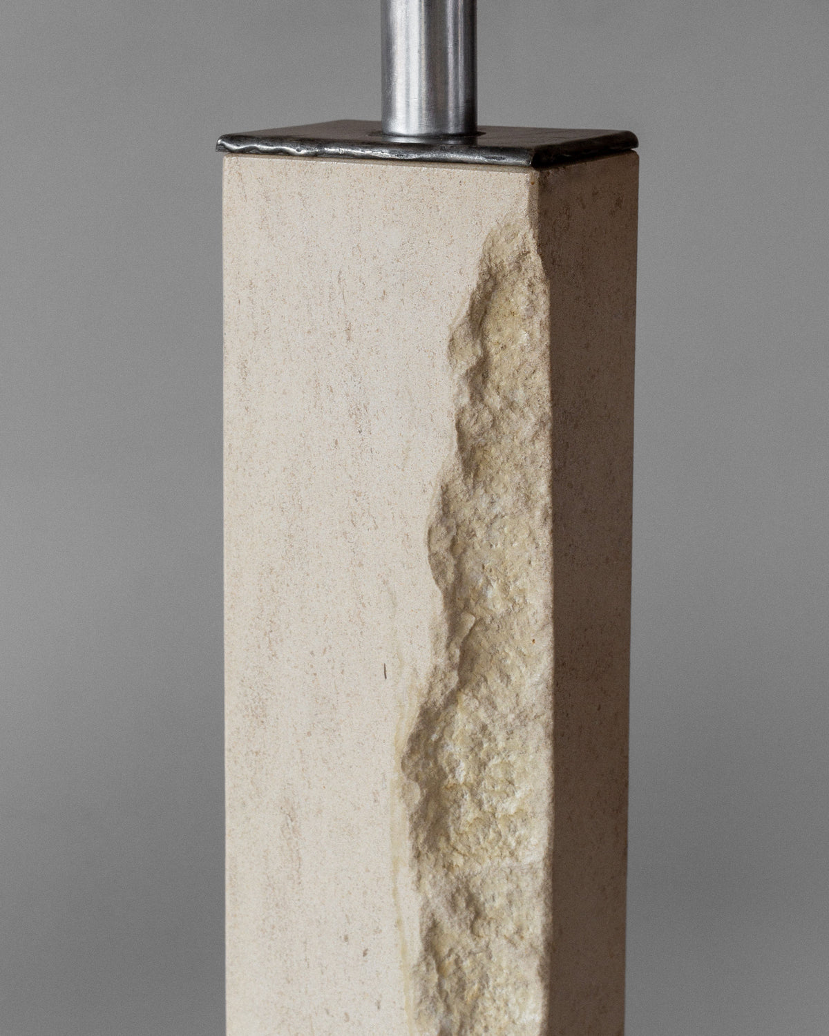 The Modernist Travertine and Metal Table Lamp by Spigel features a rectangular stone pillar with one side's rough texture contrasting against a smooth surface, topped with a sleek mid-century modern metal fixture, all set against a plain gray background.