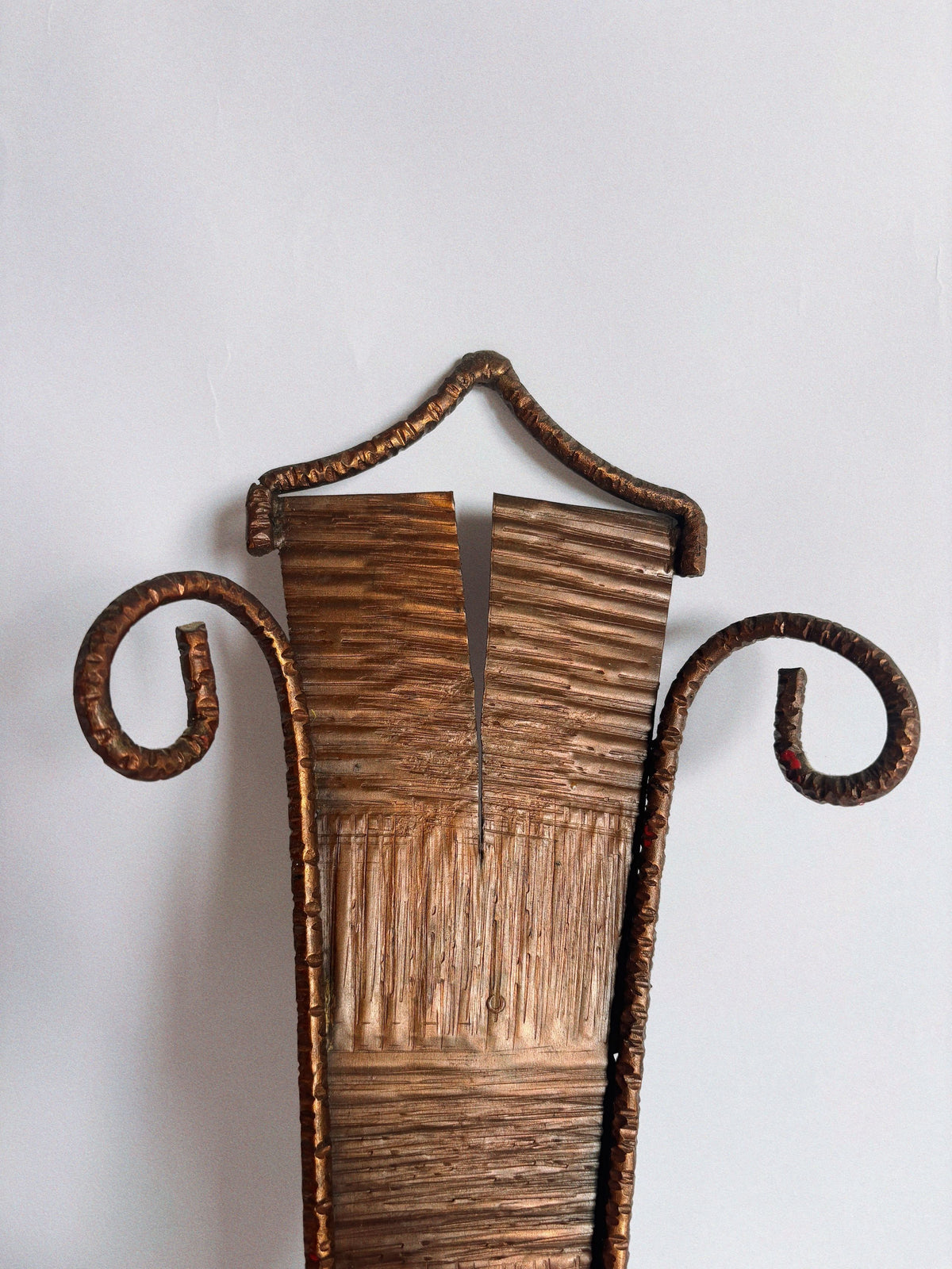 A close-up of the Huerto Metalwork Wall Candleholder shows its textured vertical body, curled side ends, and a triangular arch on top, highlighting artisan design against a plain light background.