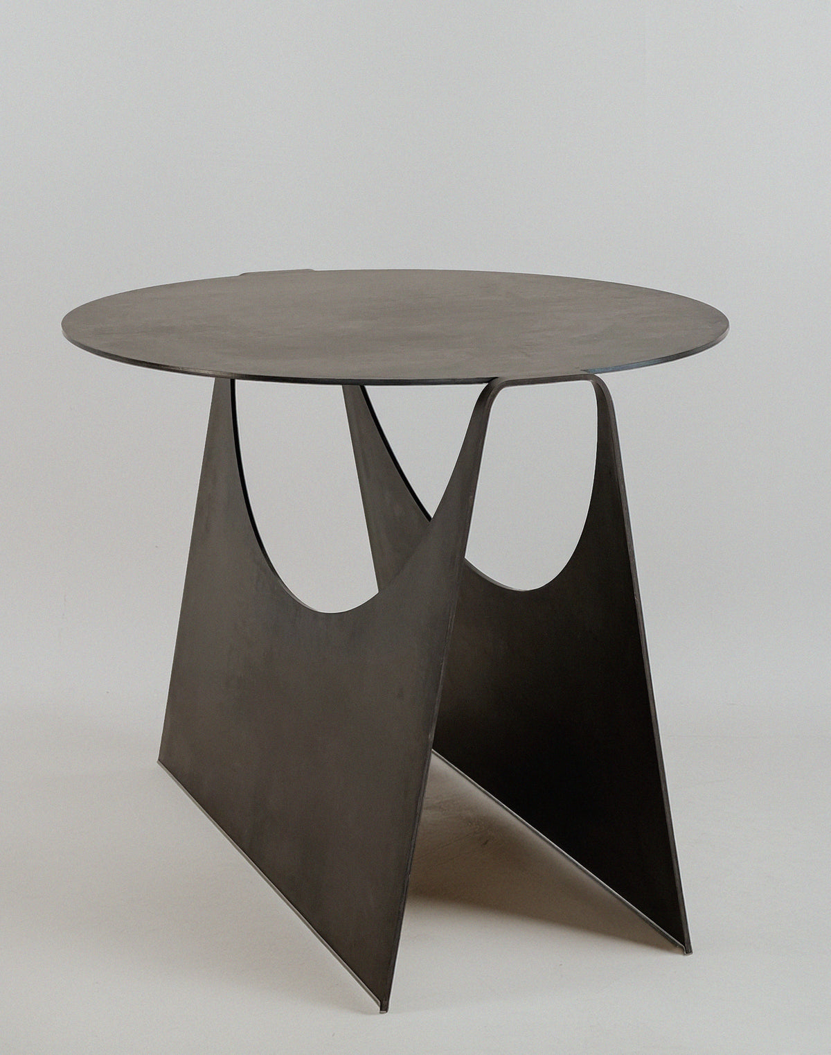 The 'Maru' Side Table by Modan features a modern, minimalist design with a round steel sheet top and two triangular supports with large semicircular cutouts, creating a distinctive look against a plain white background.