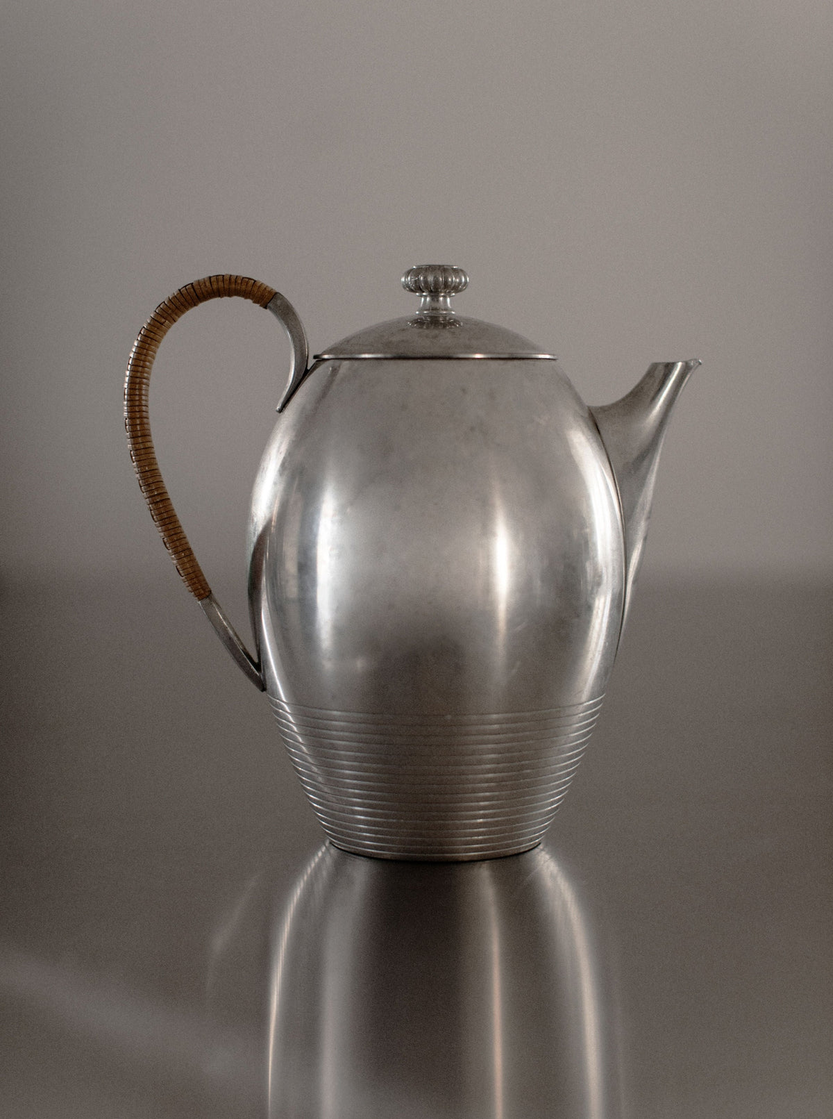 The "Art Deco Teapot" by Out For Lunch, with its sleek, metallic finish, rounded body, and distinctive ribbed lower design, sits gracefully on a reflective surface. Featuring a curved spout and a woven handle, this teapot's elegant shape is accentuated by the minimalist background, enhancing the overall aesthetic appeal of your dining experience.