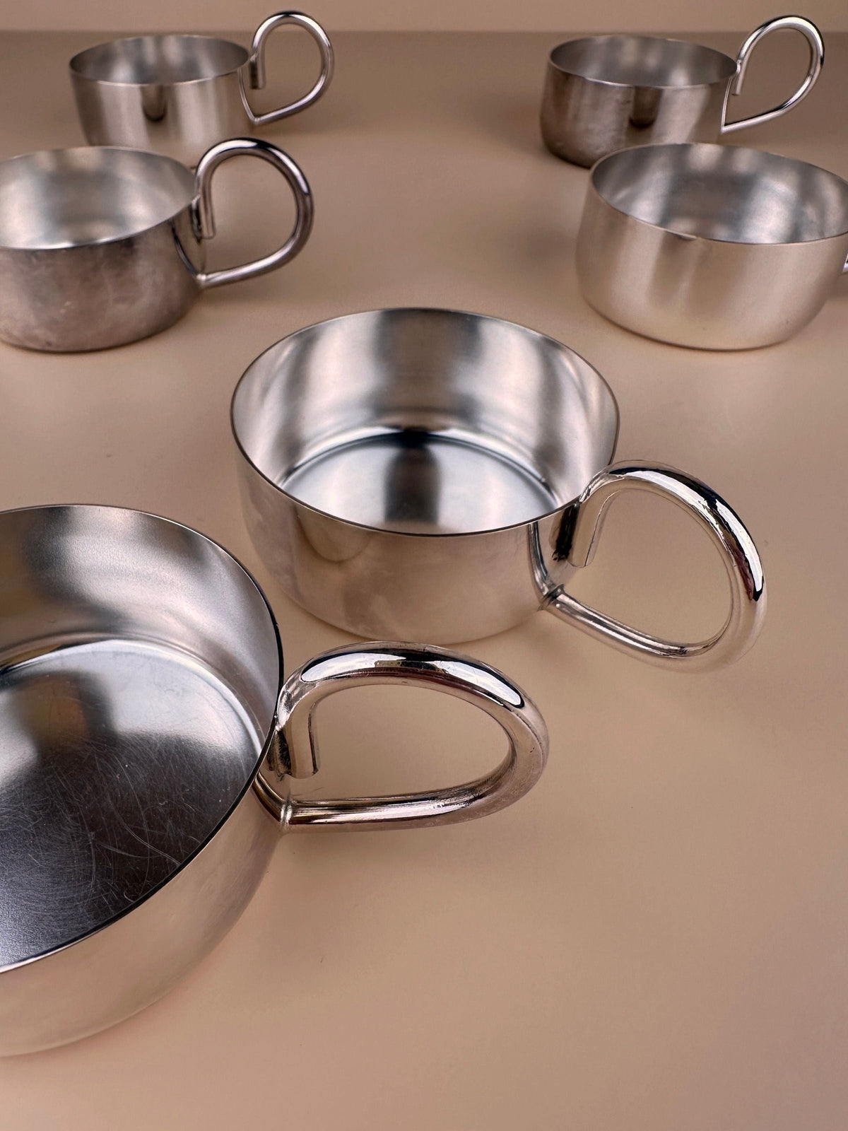 The image displays 6 swiss made stainless steel espresso cups by Dodo Vintage, arranged on a beige surface. The espresso cups have rounded handles and a shiny, reflective finish. Some are positioned upright, while others are tilted or laying on their sides, creating a varied composition.