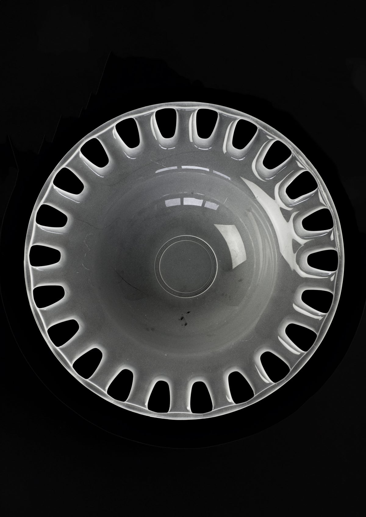 A symmetrical, round hubcap with a glossy surface and cut-out holes along the outer edge resembles the elegance of septembre studios' Large Bowl of Milk Glass, set against a sleek black background.