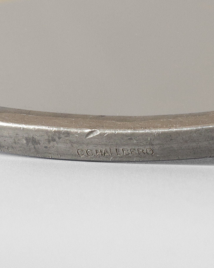 A close-up of the Art Deco Metal Mirror from Spigel reveals an inscription that reads "C.G. HALLBERG" on its surface, exuding Art Deco elegance. The focus is on the engraved text, with the rest of the mirror slightly blurred, emphasizing its vintage condition and subtly worn allure.