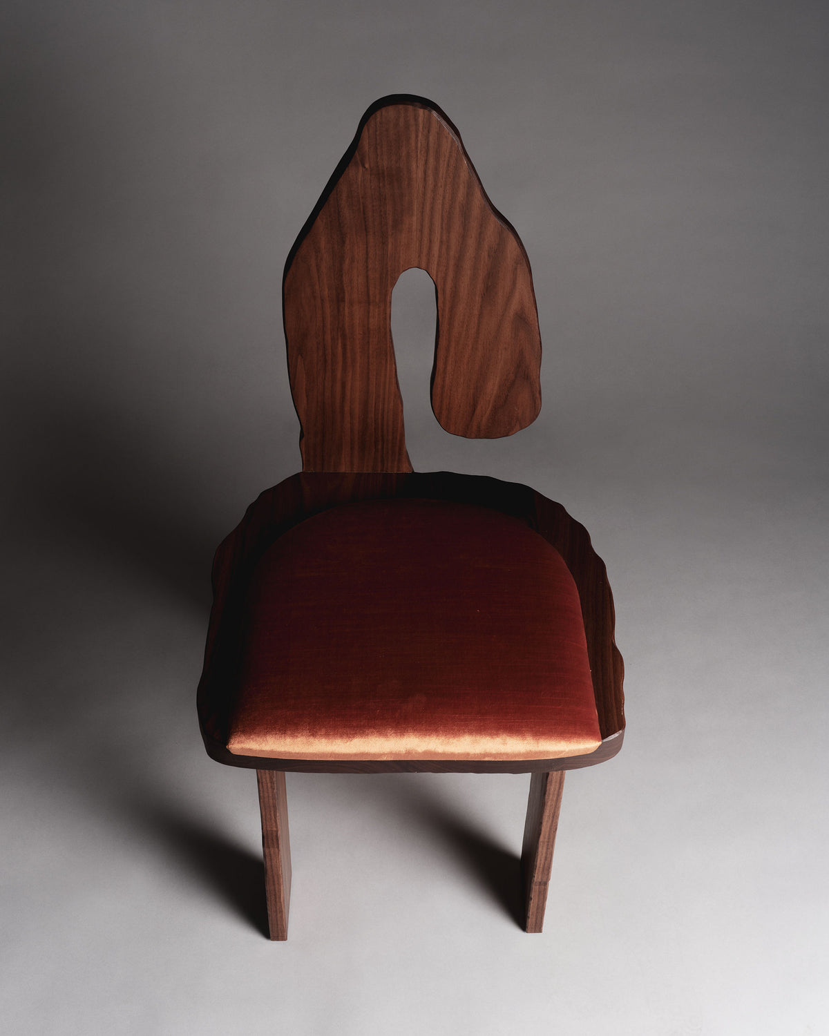 The Muisca Dining Chair by Ombia Studio has a keyhole-shaped abstract backrest. Made from oiled walnut, its seat is upholstered in rust-colored mohair, set against a neutral background.