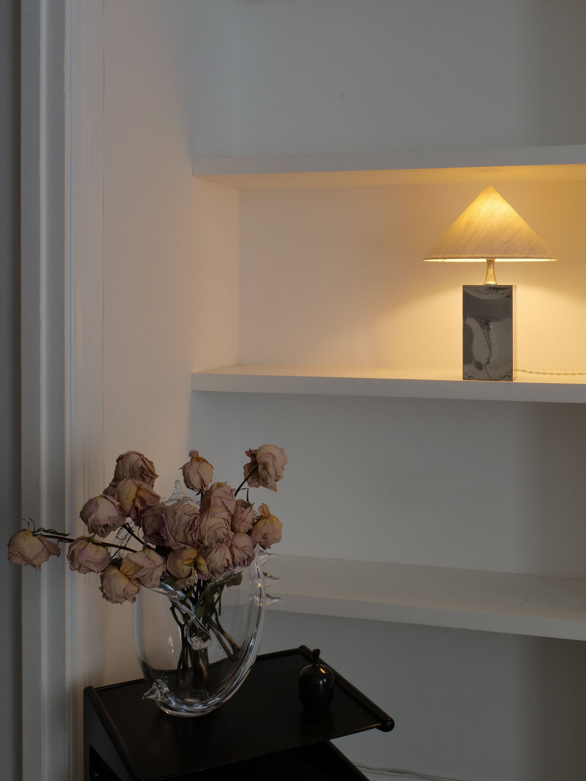 A minimalist room with white walls and built-in shelves. A small lamp with a warm yellow light sits on a middle shelf. On a black side table is a Barbier Small Box Lamp 1970s by Collection apart beside a clear vase holding wilted pink roses.