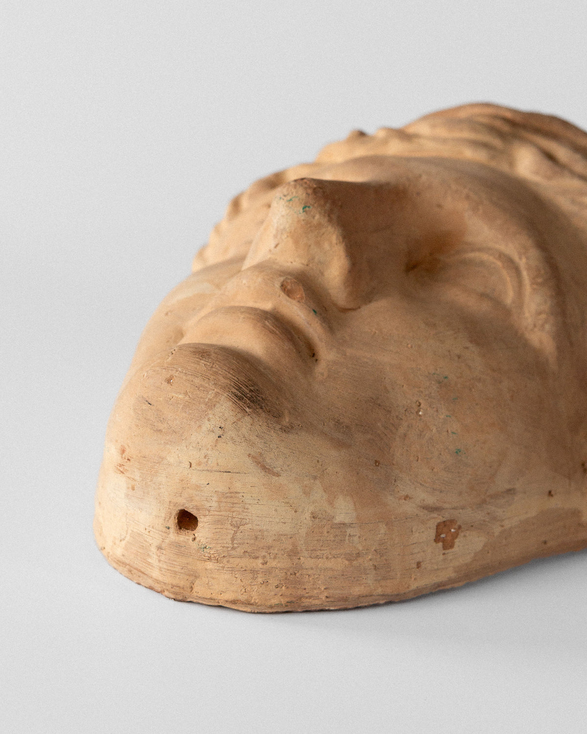 A close-up of the Spigel Terracotta Wall Mask Planter reveals a weathered, unglazed sculpture with a serene face lying on its back. The texture shows wear and small imperfections, emphasizing the neo-classical nose and lips against a plain, neutral background.