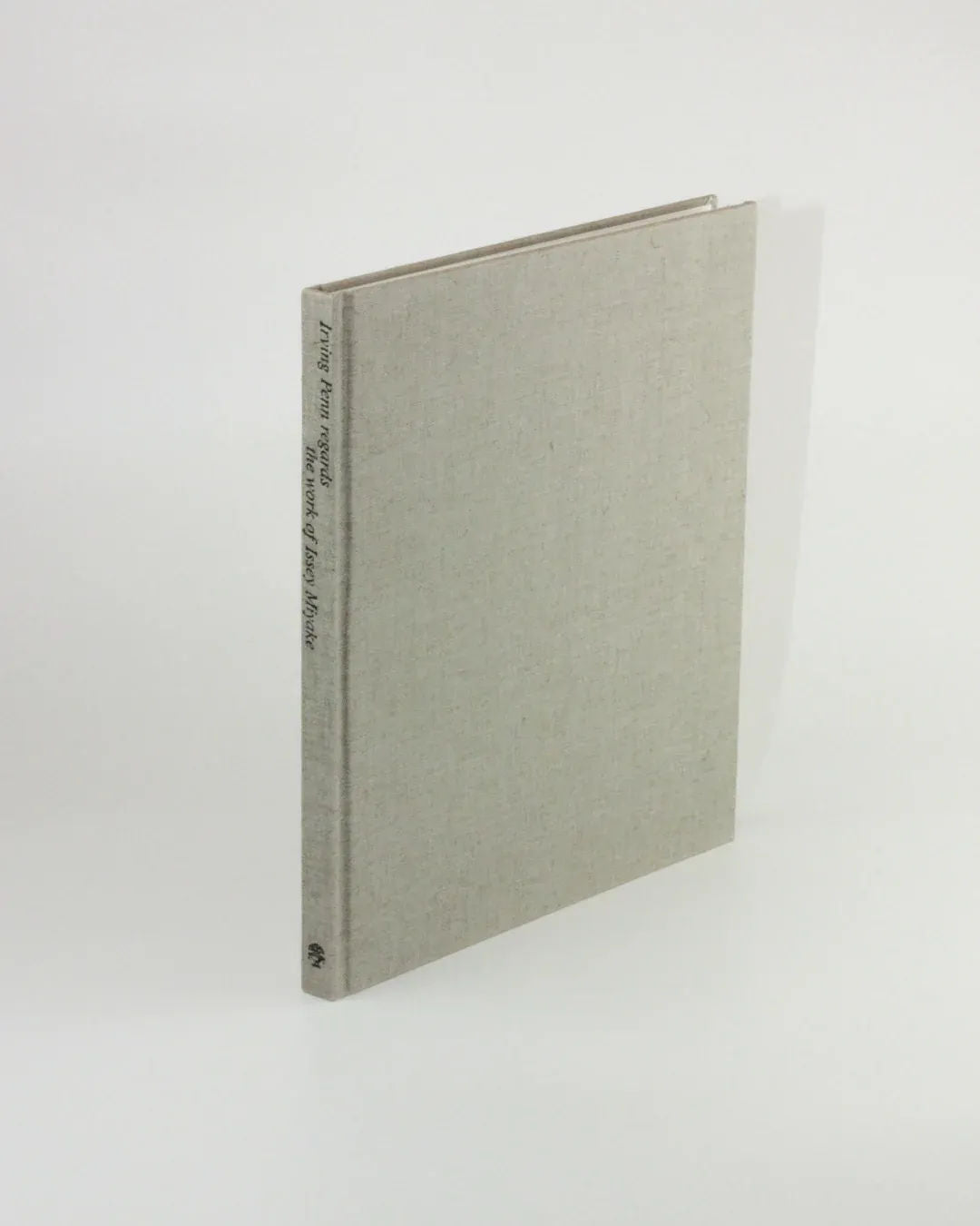 A closed, beige hardcover copy of "Irving Penn regards the work of Issey Miyake" from Boga Avante Shop stands upright against a plain white background. The spine is adorned with small, simple black text reminiscent of Irving Penn's iconic fashion photography. The book has a minimalist and clean appearance.