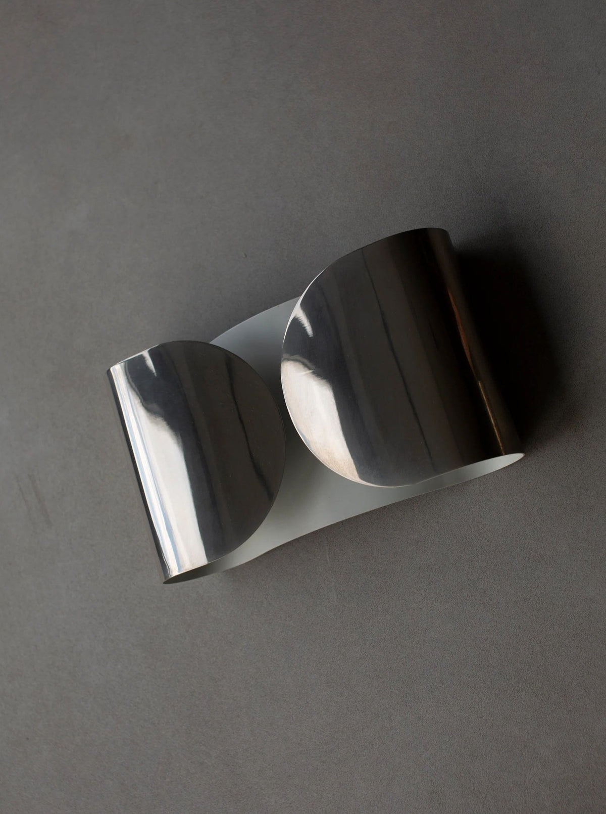 A sleek, modern metal bracelet with a wide, shiny silver band lies on a flat grey surface. Reminiscent of CU-RA-TED's elegant designs like the Chrome Wall Lamp by Tobia Scarpa 80's, the band curves into two interlocking semicircles, creating a stylish and minimalist design.