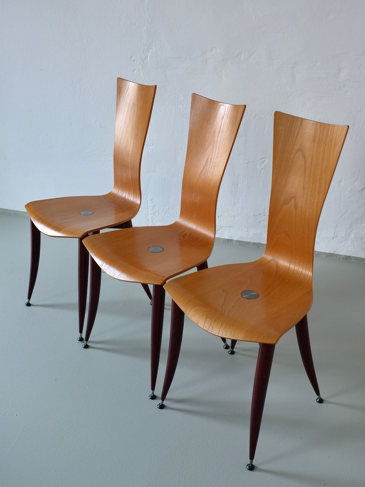 Three Veter Vintage chairs from the 1980s, featuring plywood with burgundy tapered legs and curved backs, are lined up on a light gray floor against a plain white wall. Each chair has a round metal accent on the seat.