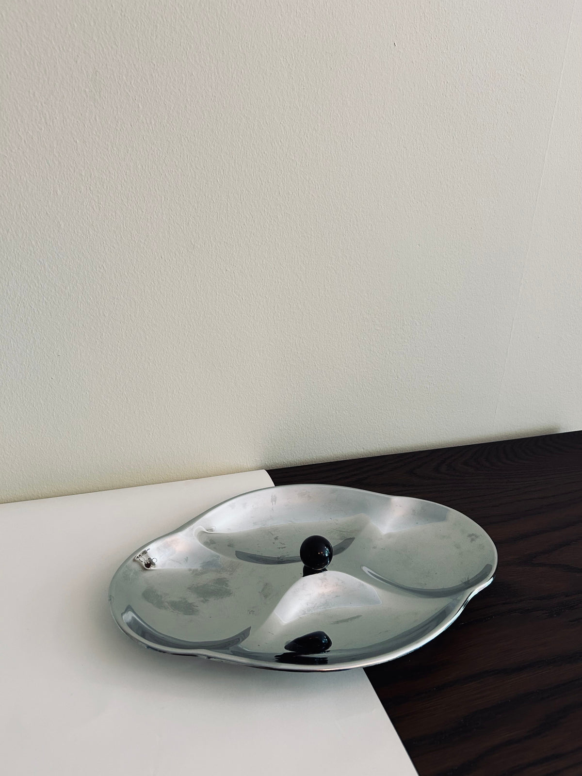 A Mid Century Serving Tray by Flock Studio, featuring curved sections and two small black objects, sits elegantly on a surface with white and dark wood patterns against a plain light wall.