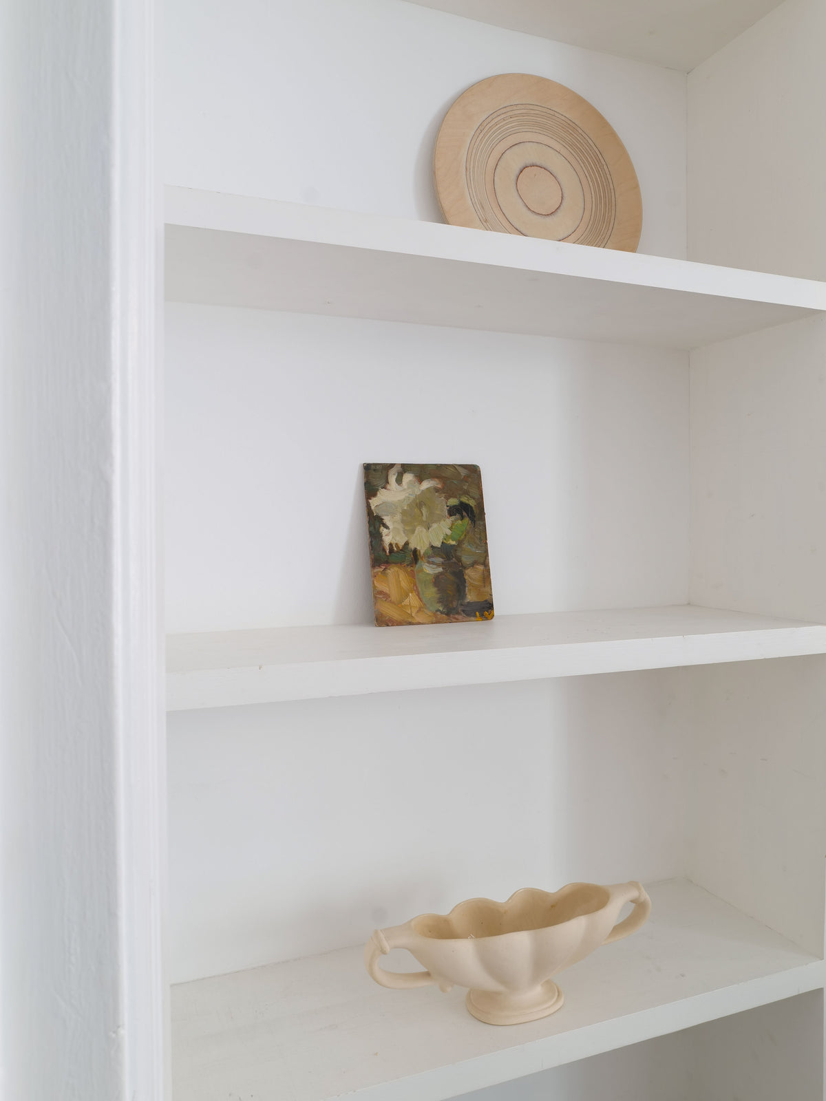 In a white, empty bookshelf, three items are displayed: a round, wooden plate with concentric rings on the top shelf, a small framed abstract painting with floral accents on the middle shelf, and on the bottom shelf sits a cream-colored ceramic bowl crafted from Devon clay and featuring two handles alongside Collection Apart's Pleated Vase 1930s.