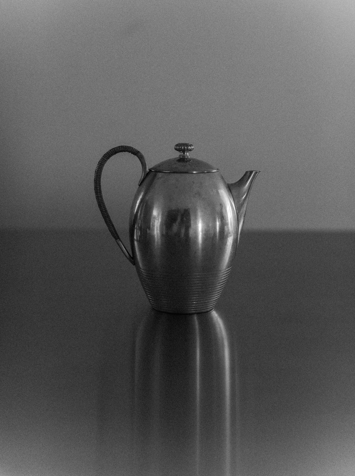 A black and white photograph of the Art Deco Teapot by Out For Lunch, featuring a pewter finish with a rounded body, a curved spout, a small knob on the lid, and a textured handle, placed on a smooth reflective surface. The image captures the aesthetic appeal that enhances any dining experience.