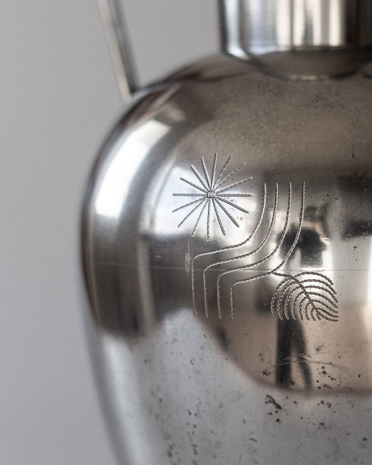 Close-up of a shiny pewter surface with engraved geometric patterns, reminiscent of Scandinavian metalwork by Rolf Engströmer. The Large Modernist Pewter Amphora by Spigel from the 1930s features starbursts and wavy lines, offering slightly distorted reflections against a neutral gray backdrop.