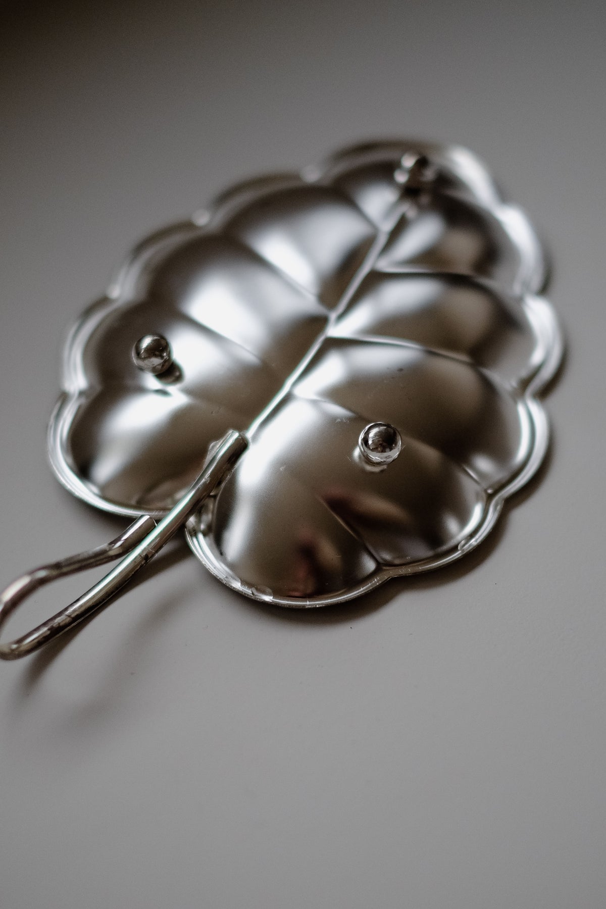 The septembre studios Serving Dish Leaf is a shiny, metallic serving dish with a textured surface and vintage charm. It features a stem-like handle and its reflective, smooth appearance is emphasized against the softly blurred background.