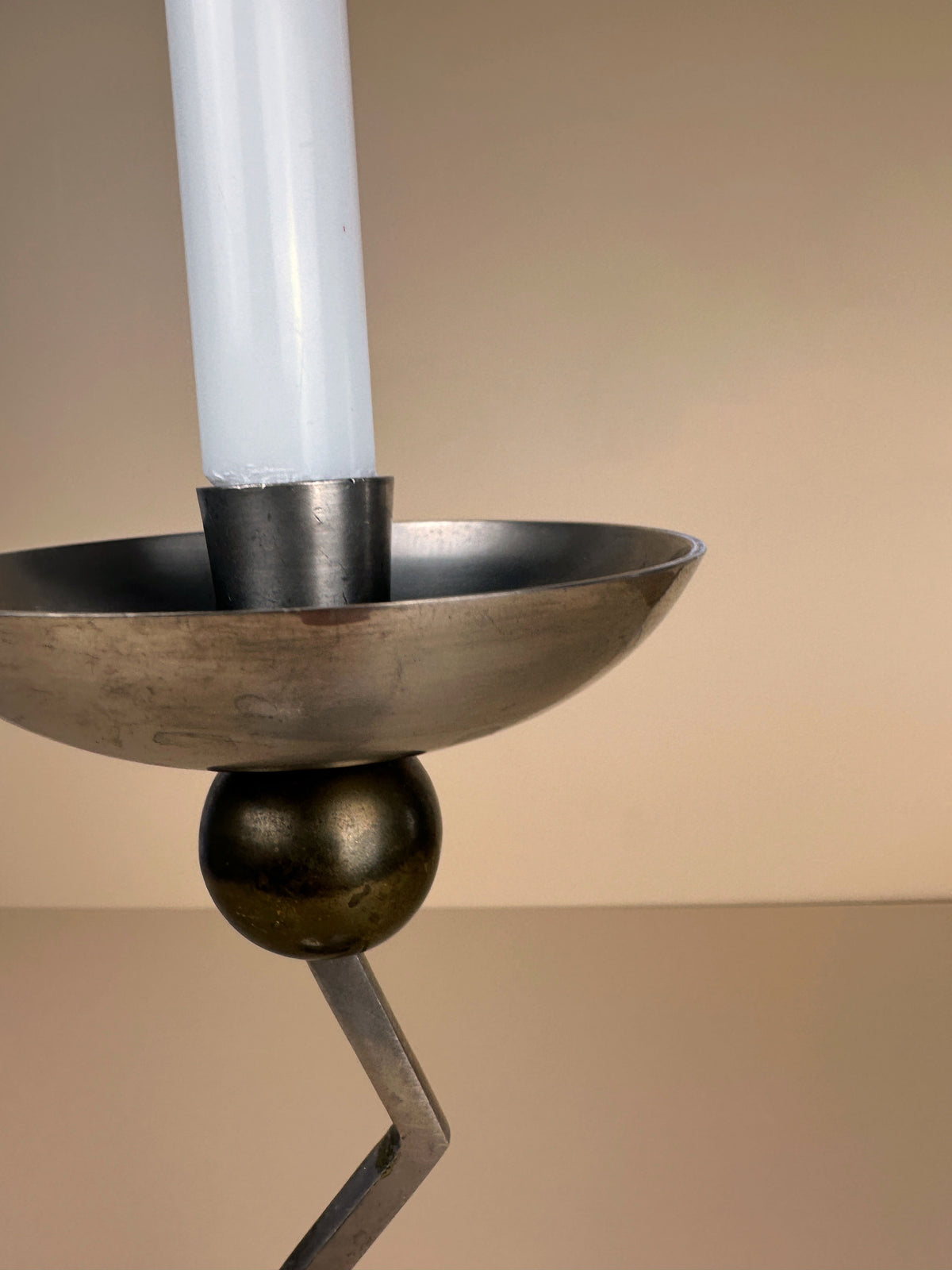 A close-up of the Postmodern Zig Zag Candle Holder by Dodo Vintage, featuring a silver-colored metal design with a white candle. The holder showcases an angular stand connected to a round base, embodying a Memphis-style aesthetic against a plain beige background.