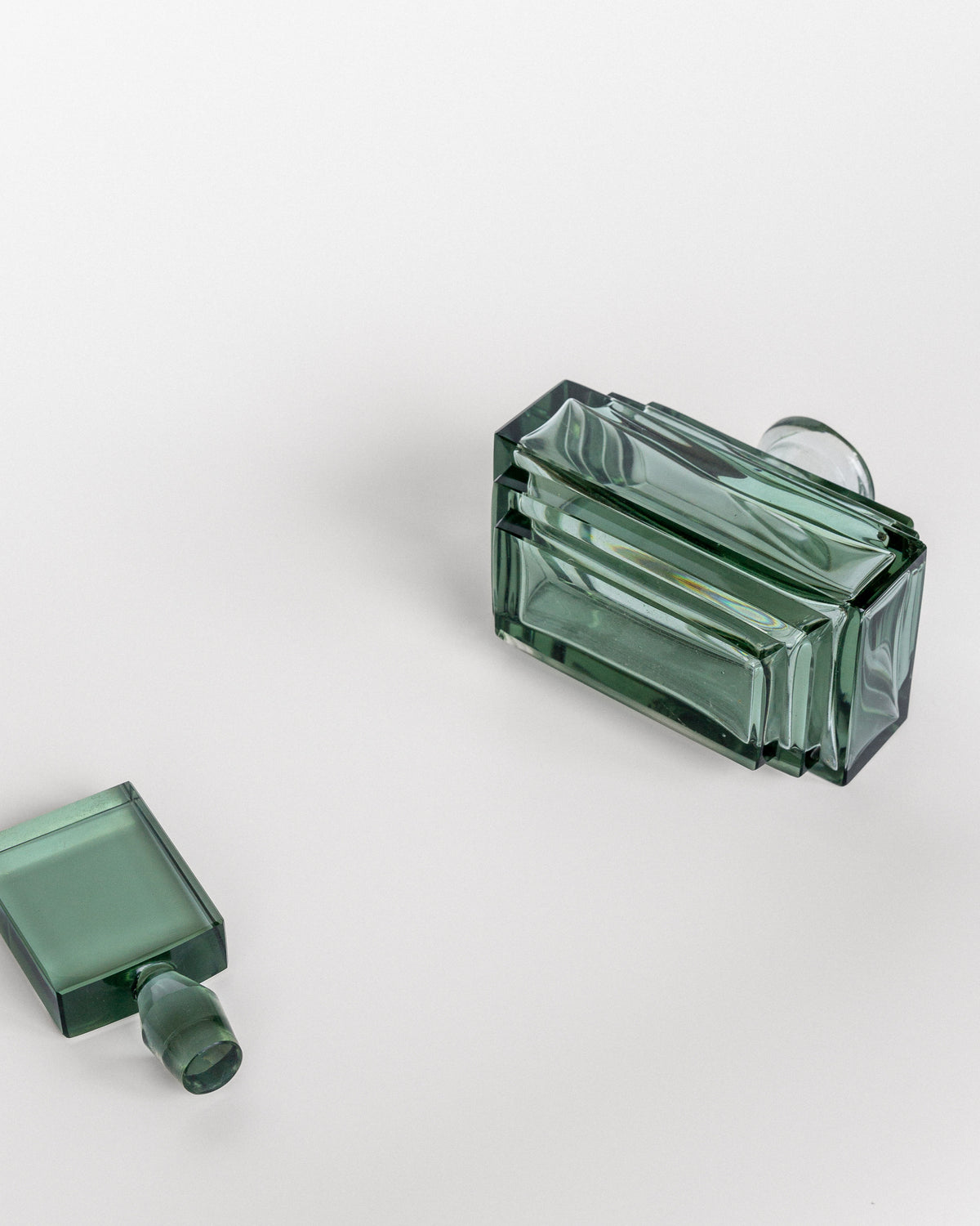 A Spigel Art Deco Perfume Bottle from the 1930s lies on its side, with its matching rectangular cap detached and resting nearby on a white surface. The green glass bottle, of Czech origin, has a minimalistic and sleek design that reflects light softly.