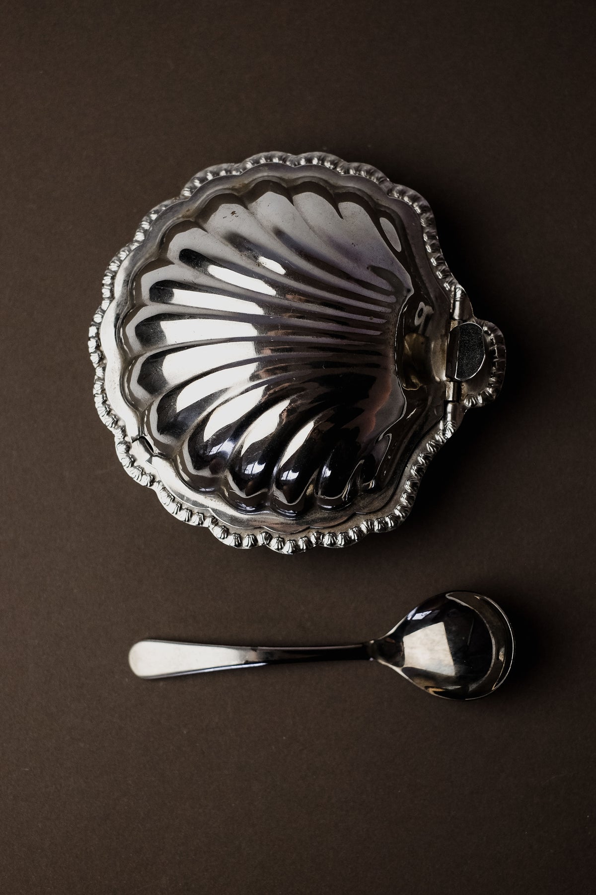 The Shell Shaped Butter Dish with Spoon by septembre studios features a vintage brass design, resembling a silver clamshell with a decorative scalloped edge. Resting on a dark surface, the included small metal spoon enhances the refined dining experience.