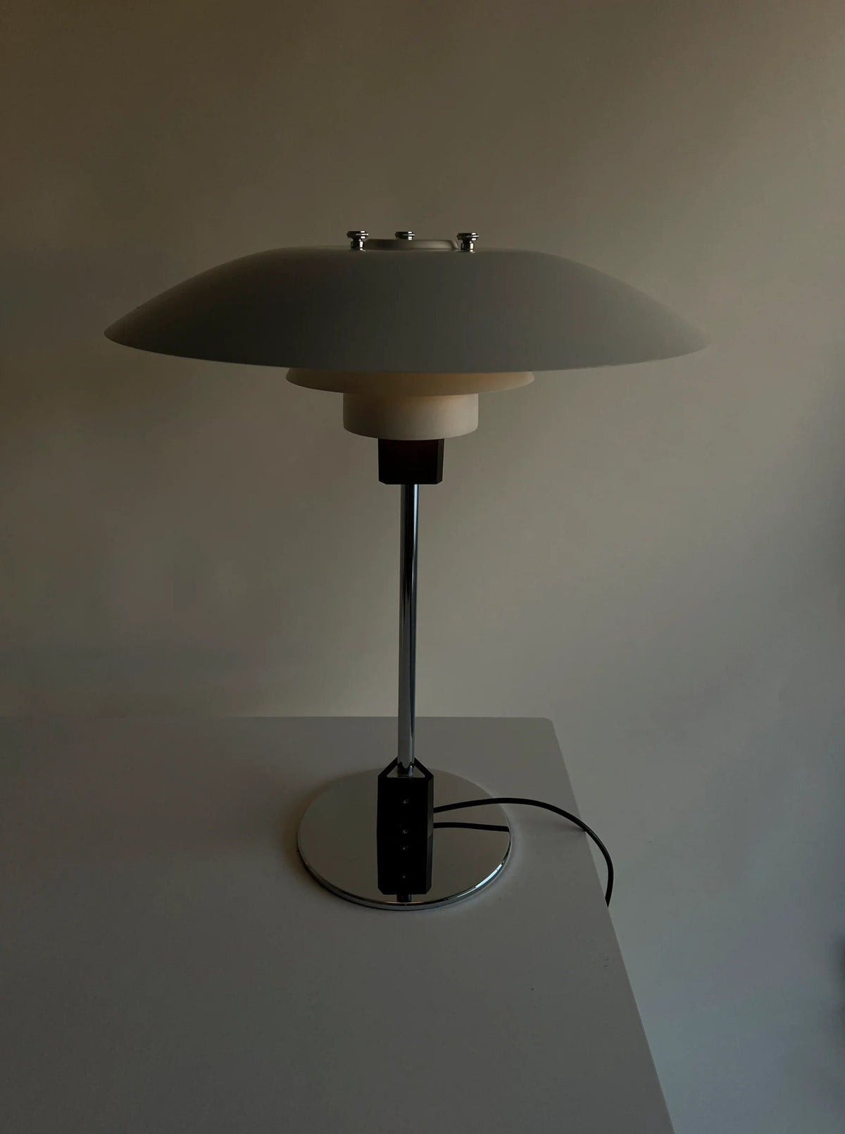 A sleek, modern table lamp with a wide, dome-shaped shade and a minimalist design stands on a round, chrome base. Reminiscent of Poul Henningsen's 1983 vintage table lamp designs, the CU-RA-TED Poulsen Table Lamp PH 4/3 1983 is placed on a plain, light-colored surface with a power cord extending from the base. Neutral background with soft lighting.