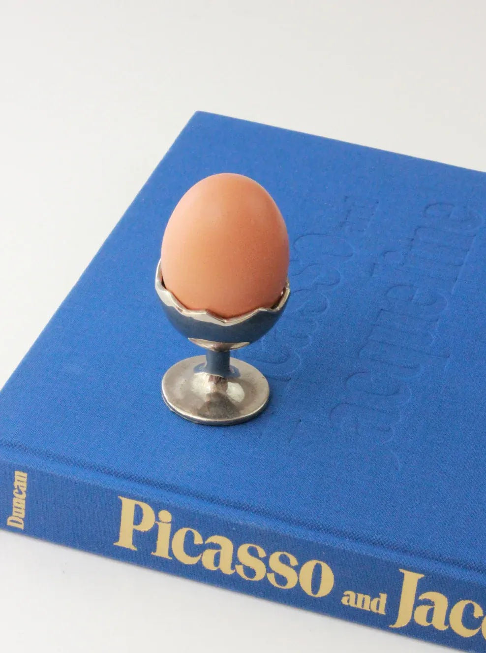 A brown egg sits in an Egg Cup by Boga Avante Shop placed on top of a blue hardcover book titled "Picasso and Jacqueline." The egg and egg cup are centered on the book, which lies on a white surface. The silver cuff of the Egg Cup by Boga Avante Shop features a decorative edge that adds a touch of elegance.