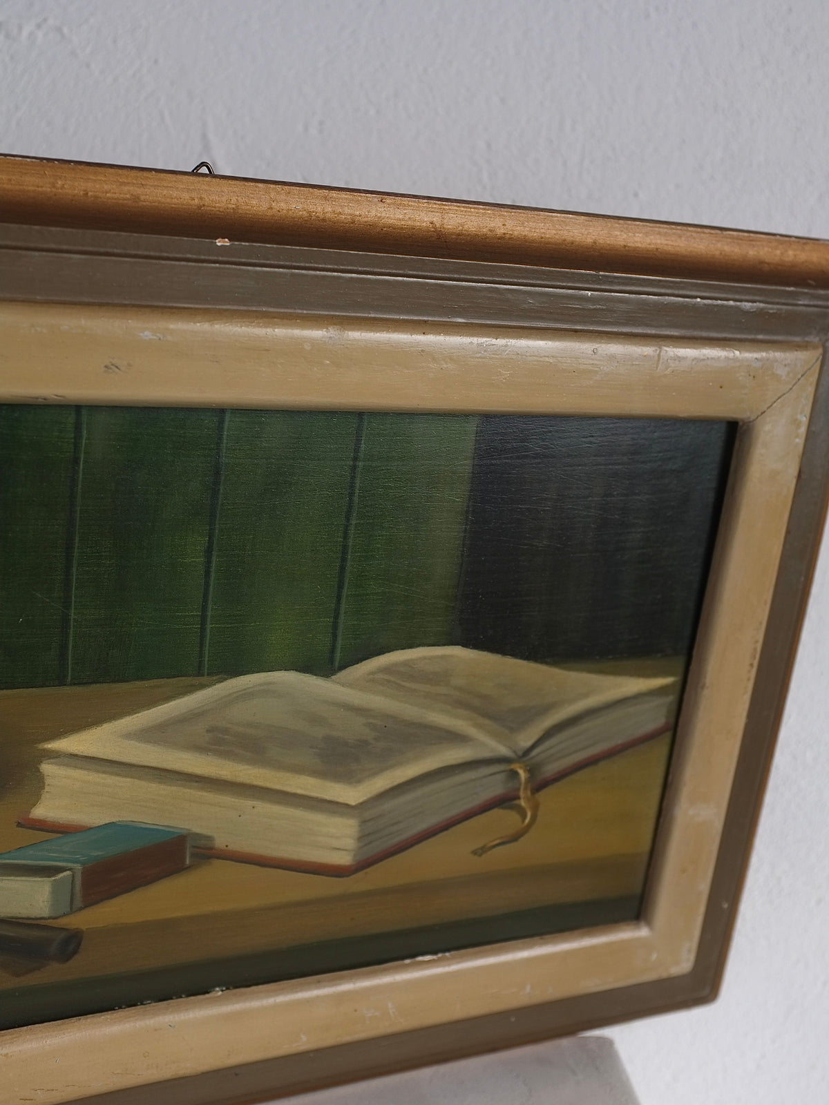 A framed painting of European origin depicting an open book lying on a wooden surface, accompanied by what appears to be a rectangular object, possibly another book or a box, in the lower left corner. This Veter Vintage Vintage Realism Still Life Oil Painting | 1970s features a green-shaded background, adding subtle contrast.
