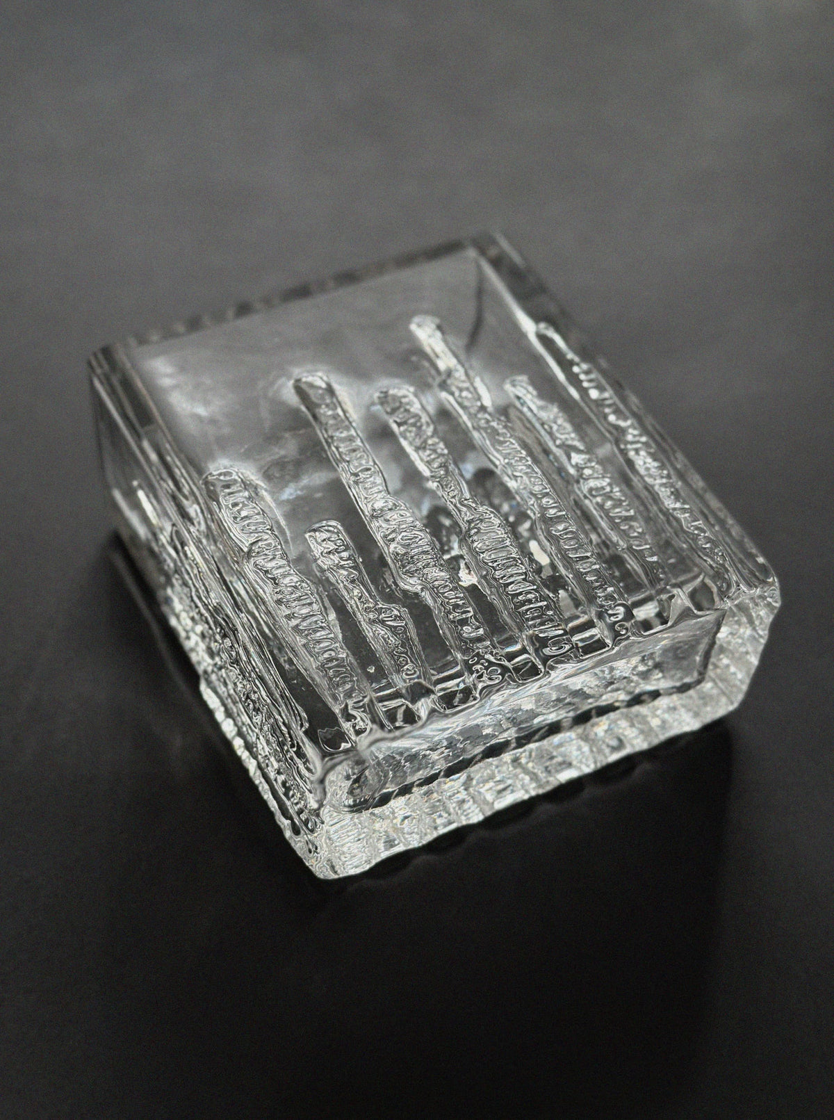 The Kosta Sweden Ice Glass Vase by Vintage Stories features a clear, rectangular minimalist design with vertical textured ridges, inspired by classic Scandinavian sculptural glass. It rests elegantly against a dark, smooth background.