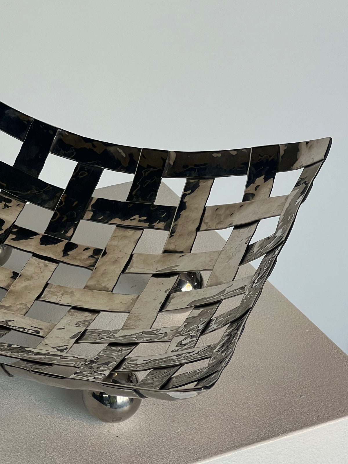 The Porter Studio Aliminum Dish on Feet features a modern metallic design with beautiful proportions and a reflective surface, set on a light pedestal against a soft white background. Its intricate weave gives it a textured and dynamic appearance.