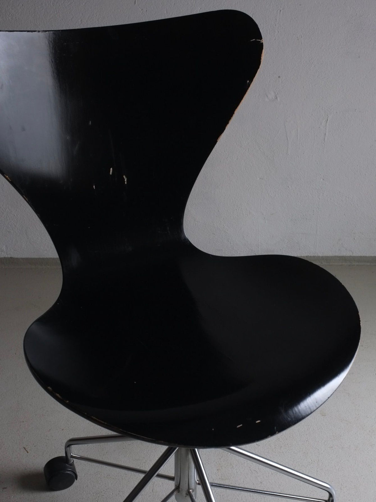 A black, modern ergonomic chair from Veter Vintage, featuring a curved backrest and slightly worn edges. It has a chrome, height-adjustable base on wheels. Positioned against a plain gray wall and placed on a neutral-colored floor, this piece evokes the elegance of the 3117 Black Swivel Chair by Arne Jacobsen for Fritz Hansen.