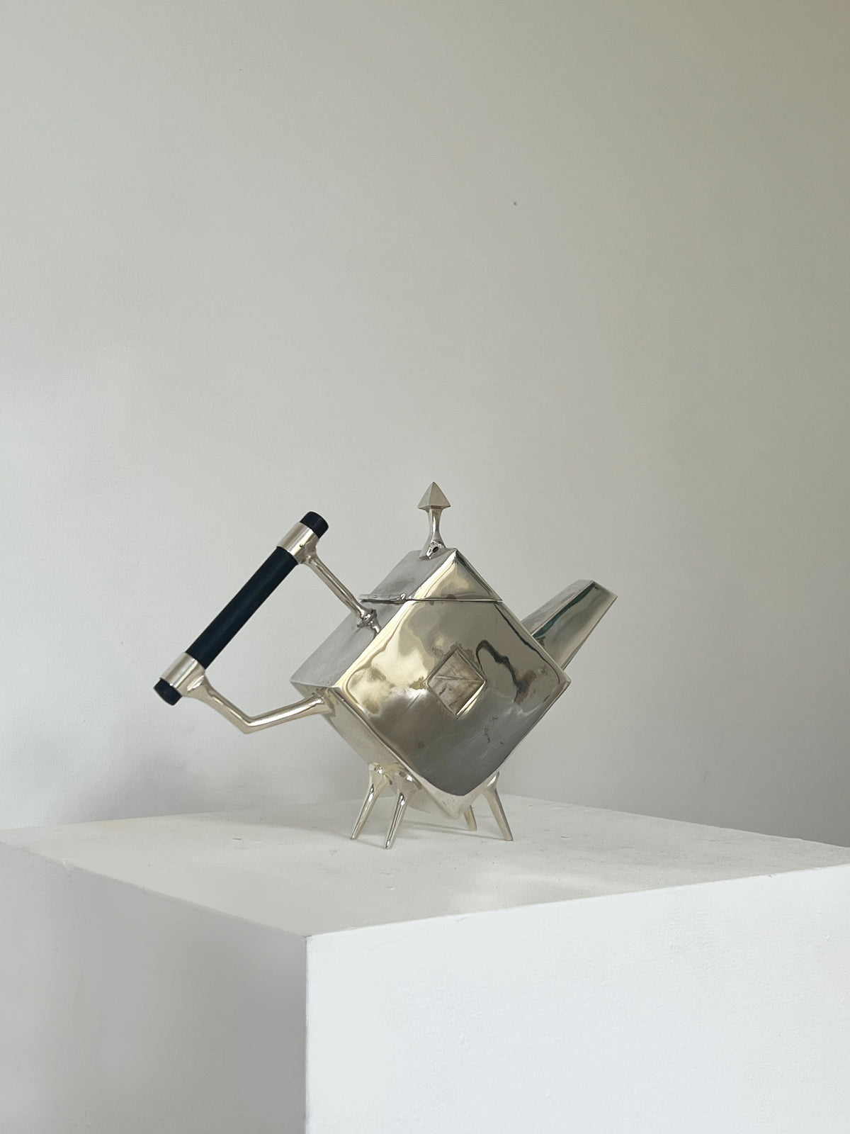 The Christopher Dresser Square Teapot by Porter Studio, featuring a geometric silver-plated design with a rectangular body, triangular spout, and black handle, rests on a white pedestal against a light-colored wall, showcasing timeless elegance.