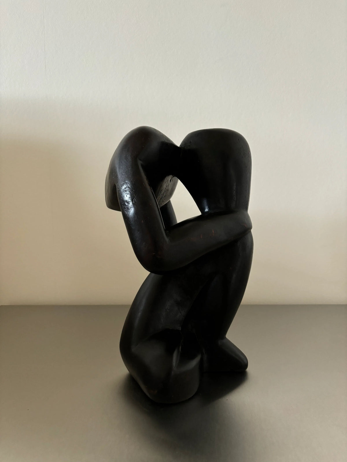 A CU-RA-TED Black Wooden Object - "The Thinker" of two faceless figures embracing, positioned on a smooth, light surface. The intertwined figures have smooth, flowing curves and minimalist features, evoking a sense of intimacy against a plain, light-colored background. The craftsmanship is evident in its sleek black finish.